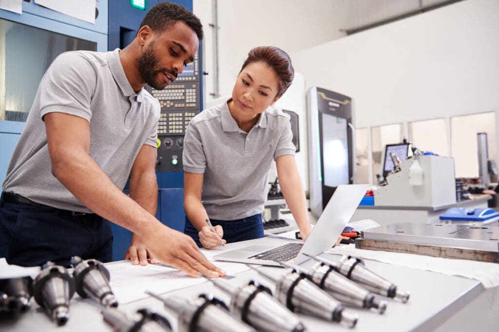 Two engineers review plans on a table in a workshop, surrounded by machinery and tools, emphasizing collaboration in manufacturing.