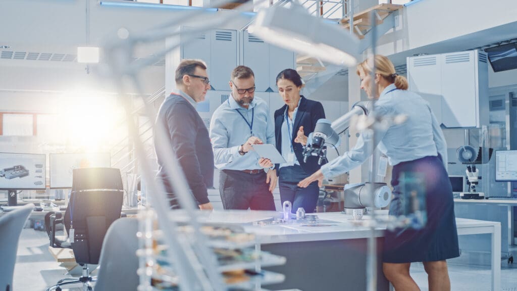 A team of four professionals discuss a project in a modern laboratory, examining a tablet and working with robotic equipment.