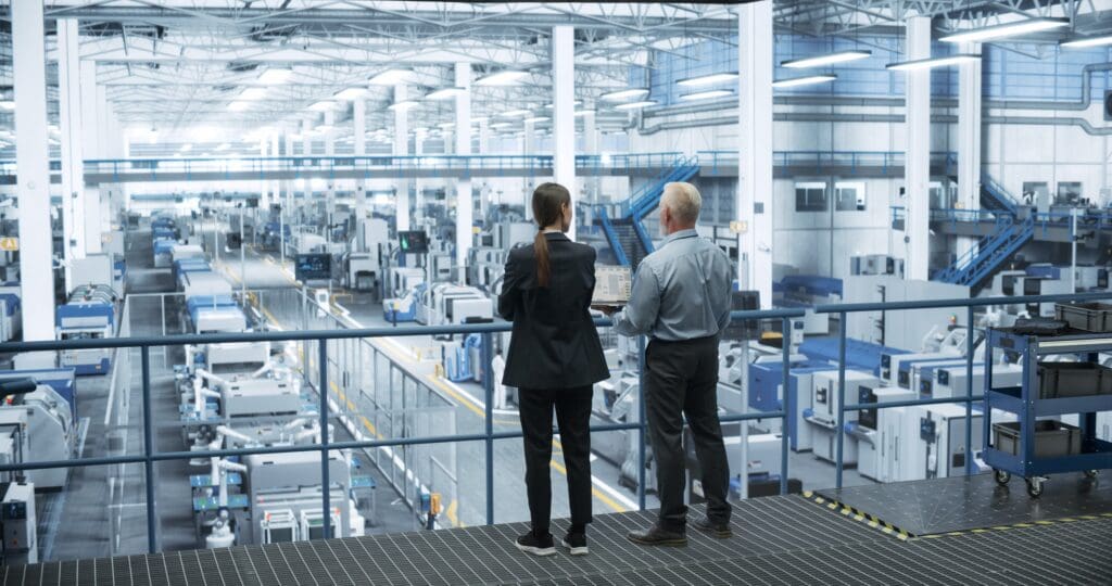 Two professionals observe a modern manufacturing facility from a balcony, showcasing advanced machinery and production lines below.