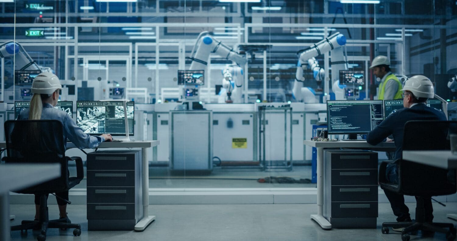 Two engineers monitor robotic systems in a high-tech factory. Screens display data as automated arms work in the background, showcasing advanced automation.